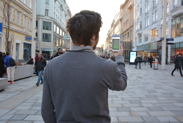 Person von hinten, die vor einer Fußgängerzone steht und ein Smartphone in die Höhe hält.