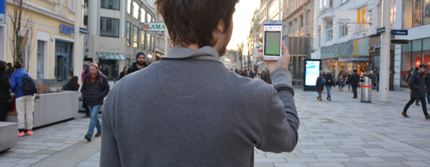 Person von hinten, die vor einer Fußgängerzone steht und ein Smartphone in die Höhe hält.