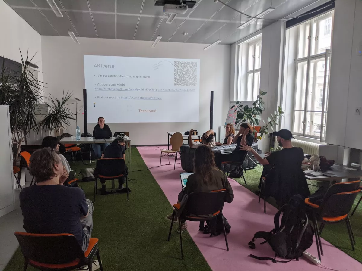 Image of a presentation in a white classroom with green and pink carpet. There are 6 students and two instructors in the room, one of the instructors is sitting at a desk doing the presentation.