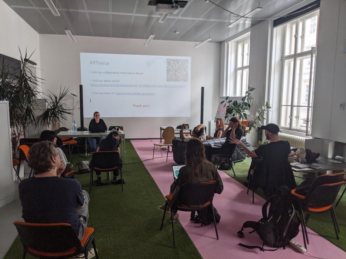 Image of a presentation in a white classroom with green and pink carpet. There are 6 students and two instructors in the room, one of the instructors is sitting at a desk doing the presentation. 